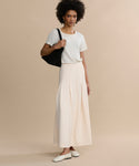 A woman stands against a beige background wearing a white short-sleeve t-shirt, the Florence Skirt in cream crepe, white flats, and carrying a black shoulder bag.