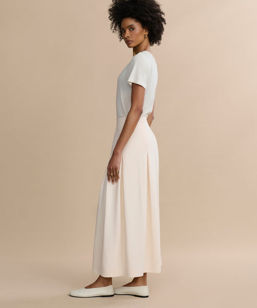 A woman with short curly hair stands sideways against a beige background, wearing a white short-sleeve t-shirt, white slip-on shoes, and the Florence Skirt—a long cream crepe skirt.
