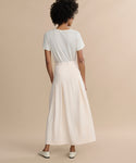 Facing away, a person with short curly hair wears the Florence Skirt—a high-waisted, cream crepe midi skirt with pleats—paired with a short-sleeve white t-shirt and white shoes against a plain beige background.