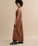 A person with short curly hair stands sideways, wearing the Florence Slip Dress with a fitted bodice and thin straps, made from crepe fabric, paired with black ankle boots against a beige background.
