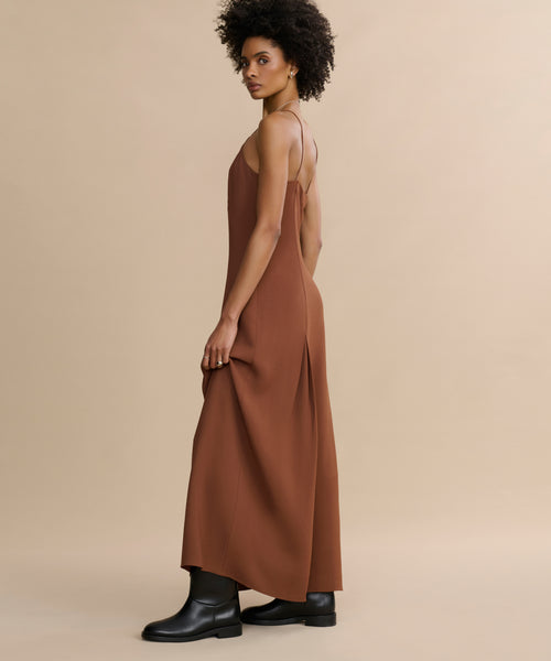 A person with short curly hair stands sideways, wearing the Florence Slip Dress with a fitted bodice and thin straps, made from crepe fabric, paired with black ankle boots against a beige background.