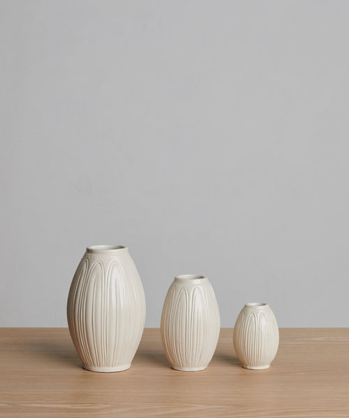 Three cream-colored Porta Ceramic Vases of different sizes, featuring vertical ridges, are arranged in a row on a light wooden surface with a plain gray background.