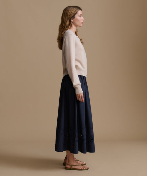 Woman wearing an oat cashmere cardigan with a blue skirt against a beige wall. 