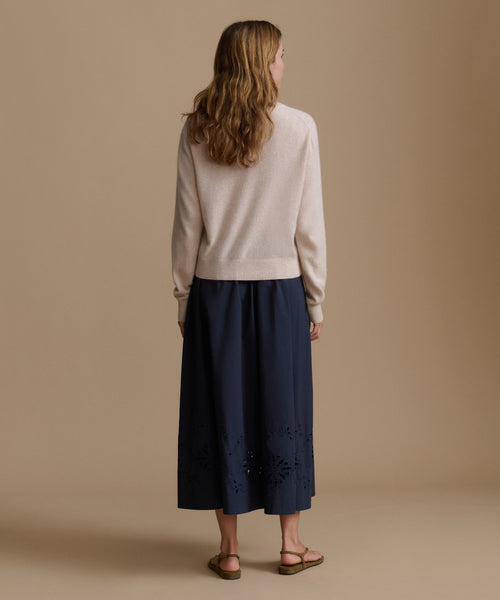 Woman wearing an oat cashmere cardigan with a blue skirt against a beige wall. 