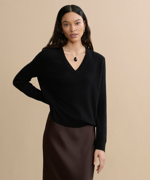 A woman with long dark hair wears the Flynn Cashmere Sweater in black, paired with a brown skirt and pendant necklace, standing against a plain beige background with one hand on her hip.