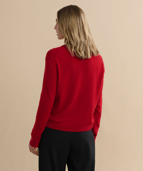 A person with shoulder-length blonde hair, wearing the Flynn Cashmere Sweater in bright red and black pants, stands facing away from the camera against a plain beige background.