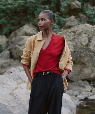 Standing outdoors before rocks and greenery, a person wears a tan jacket over the Flynn Cashmere Sweater—a relaxed fit knit v-neck—paired with black pants, hands in pockets, gazing to the side.