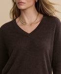 Wearing the Flynn Cashmere Sweater in dark brown and layered gold necklaces, a woman with wavy blonde hair and stud earrings is shown from the shoulders up against a beige background.