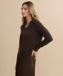 A woman with wavy blonde hair wears the Flynn Cashmere Sweater in dark brown and a brown pleated skirt. She stands in profile against a beige background, one hand on her hip.