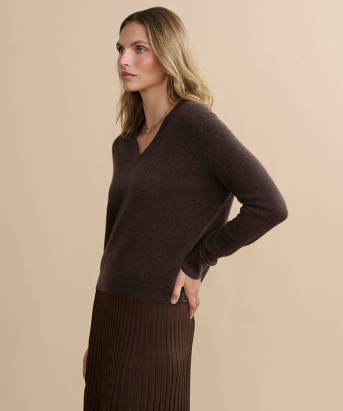 A woman with wavy blonde hair wears the Flynn Cashmere Sweater in dark brown and a brown pleated skirt. She stands in profile against a beige background, one hand on her hip.