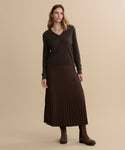 A woman with wavy, shoulder-length hair stands against a beige background wearing the Flynn Cashmere Sweater in dark brown, a matching pleated midi skirt, brown suede boots, minimal jewelry, and a neutral expression.