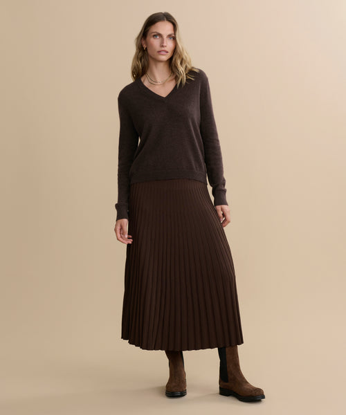 A woman with wavy, shoulder-length hair stands against a beige background wearing the Flynn Cashmere Sweater in dark brown, a matching pleated midi skirt, brown suede boots, minimal jewelry, and a neutral expression.