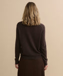 A woman with wavy blonde hair, wearing the Flynn Cashmere Sweater in dark brown and a pleated brown skirt, stands facing away from the camera against a beige background.