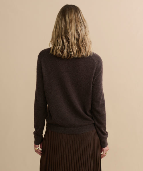 A woman with wavy blonde hair, wearing the Flynn Cashmere Sweater in dark brown and a pleated brown skirt, stands facing away from the camera against a beige background.