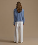 Woman wearing a blue cashmere sweater and white pants against a beige background