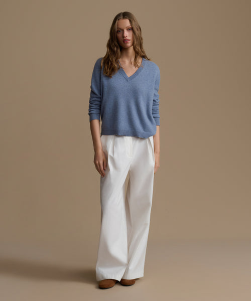 Woman wearing a blue cashmere sweater and white pants against a beige background