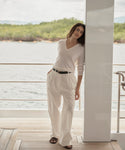 A woman stands on a wooden deck beside a white column, wearing a cream V-neck knit sweater and matching cream wide-leg trousers with a thin black belt. The background shows water and green shoreline under a bright sky.