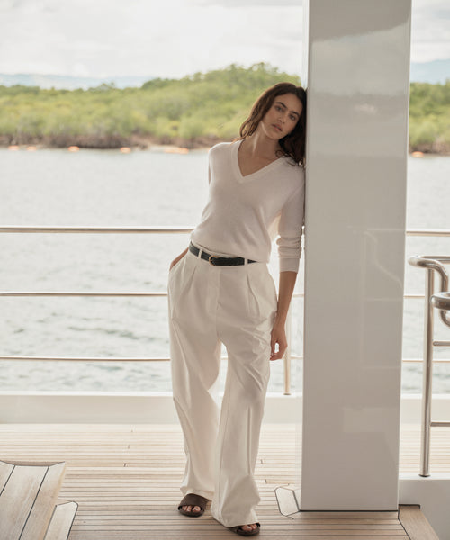 A woman stands on a wooden deck beside a white column, wearing a cream V-neck knit sweater and matching cream wide-leg trousers with a thin black belt. The background shows water and green shoreline under a bright sky.