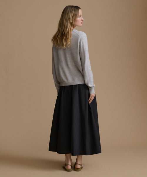 A woman is shown from the back wearing a light gray knit sweater and a long black skirt, standing against a solid tan studio background.