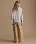 Woman wearing a white sweater and beige pants against a brown background