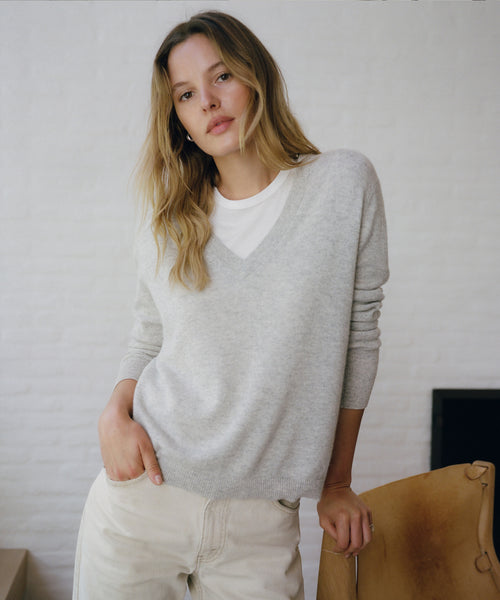 A woman wearing a light gray v-neck knit sweater over a white t-shirt and cream-colored pants, leaning against a brown leather chair in front of a white brick wall.