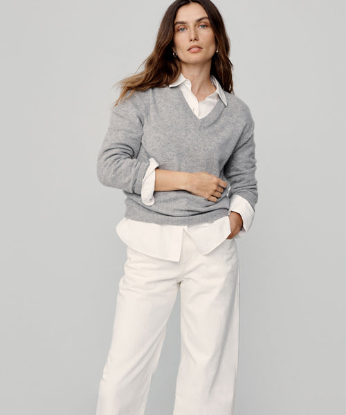 A woman stands against a plain background, wearing the Flynn Cashmere Sweater in light gray over a white collared shirt and wide-leg white pants. She has long brown hair, a neutral expression, and one hand in her pocket.