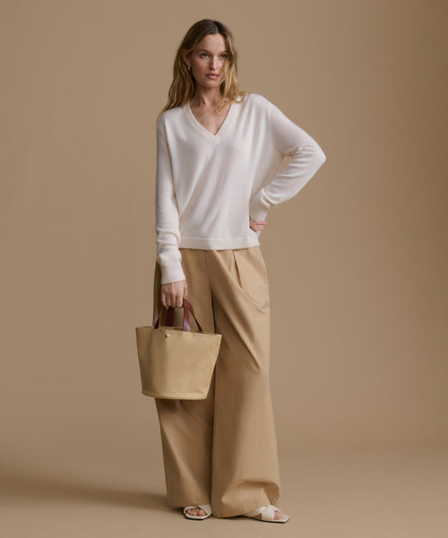 Woman wearing a white sweater and beige pants, holding a beige handbag against a brown background