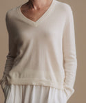 A close-up of a person wearing a cream-colored V-neck knit sweater with long sleeves and a ribbed hem, paired with a matching cream skirt against a solid beige background.