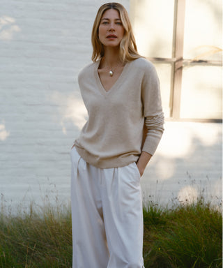 A woman with light brown hair stands outside in front of a white brick wall and window, wearing the beige Flynn Cashmere Sweater, white wide-leg pants, and a pendant necklace, with her hands in her pockets.