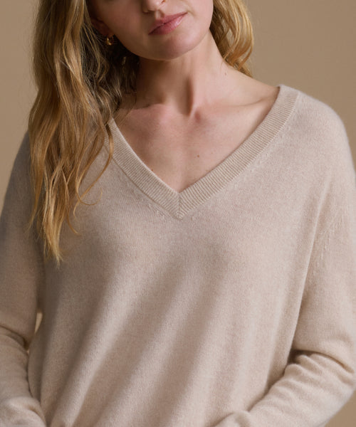 A close-up of a woman wearing a soft, light beige V-neck knit sweater against a warm beige background.