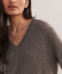 A woman with long dark hair wears the Flynn Cashmere Sweater in grey and small gold earrings, shown from the shoulders up against a neutral background.