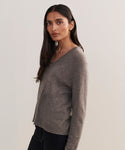 A woman with long dark hair stands against a plain light background, wearing the Flynn Cashmere Sweater—a relaxed fit knit V-neck—paired with black pants, looking slightly to the side with a neutral expression.