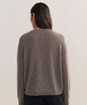 A person with long dark hair, seen from behind, wears the Flynn Cashmere Sweater in a relaxed fit grayish-brown knit with dark pants against a plain light background.