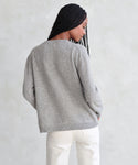 A person with long braids stands with their back to the camera, wearing the Flynn Cashmere Sweater in light gray and off-white pants against a light, textured background.
