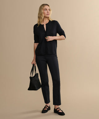 A woman with blonde hair stands against a beige background, wearing the Forme Pant, a black long-sleeve top, black shoes, and holding a black tote bag in her left hand while looking slightly to the side.