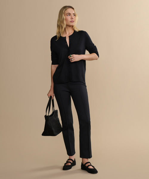 A woman with blonde hair stands against a beige background, wearing the Forme Pant, a black long-sleeve top, black shoes, and holding a black tote bag in her left hand while looking slightly to the side.