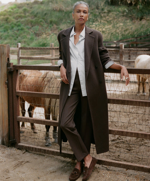 A person wearing Forme Pant, a long dark brown coat, and a white shirt stands by a wooden fence in front of horses, with greenery in the background.