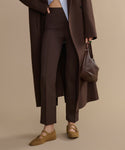 A person wearing Forme Pant, a long brown coat, tan suede Mary Jane shoes, and holding a brown handbag stands against a neutral beige background.