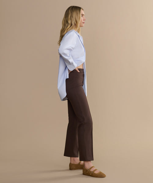 A woman with long, wavy blond hair stands in profile against a beige background, wearing the Forme Pant in a brown flared style, a light blue button-up shirt, and tan loafers, with one hand in her pocket.