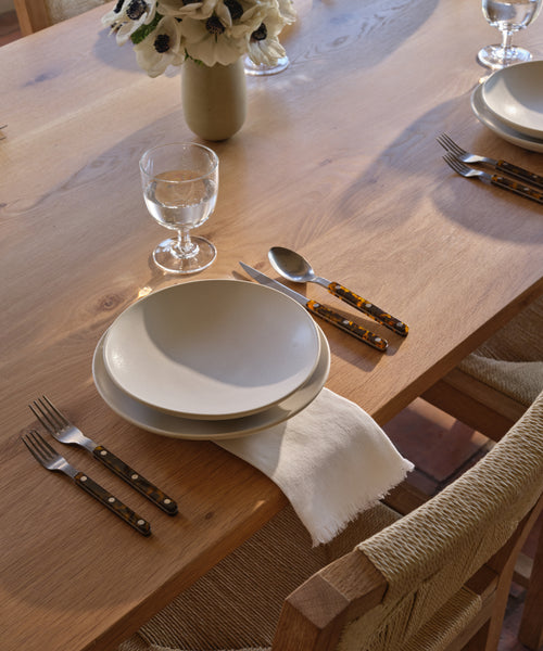 A wooden dining table set with white plates, a neatly folded Frayed Linen Napkin, a glass of water, and dark-handled cutlery. A vase of white flowers sits in the background as natural light brightens the scene.