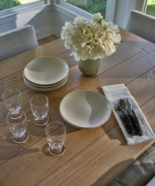 A wooden dining table set with white plates, clear glasses, black-handled cutlery on a Frayed Linen Napkin, and a vase of white flowers in a bright room with large windows.