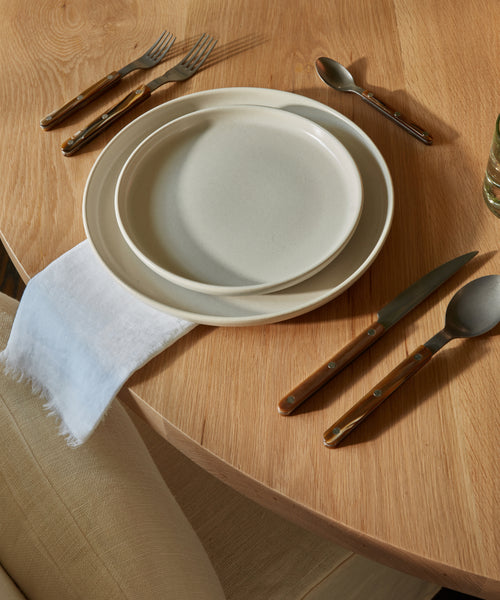A wooden dining table set with two stacked cream-colored plates, a Frayed Linen Napkin, a fork and spoon above the plates, a knife and spoon to the right, and part of a beige chair visible in the lower left corner.
