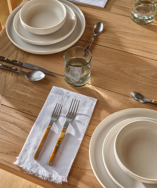 A wooden table is neatly set with cream plates and bowls, silver cutlery with brown handles, glasses of water, and a Frayed Linen Napkin in natural sunlight.