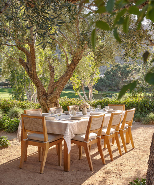 A wooden outdoor dining table with eight chairs is set under a leafy tree in a sunny garden, featuring plates, glasses, and the Frayed Linen Tablecloth with natural stripes and frayed edges, surrounded by greenery and mountain views.