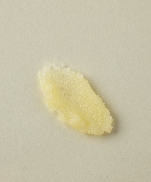 A small, irregular mound of pale yellow, grainy paste from the Travel Fresh Start Set—ideal as a body scrub—sits on a plain light beige surface.