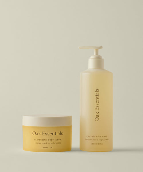 The Fresh Start Set features two yellow Oak Essentials products on a beige background: the Perfecting Body Scrub in a round jar for exfoliating (left) and the Awaken Body Wash in a pump bottle for cleansing (right).