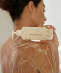 A woman with her back to the camera holds a bottle from the Fresh Start Set. Her shoulder and upper back are covered in a lather of soap as she begins to cleanse and hydrate her skin.