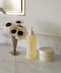A vase of white flowers and the Oak Essentials Fresh Start Set—a pump bottle to cleanse and a jar to hydrate—sit on a marble bathroom counter beneath a mirror.