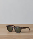 The Broadway Sunglasses by Garrett Leight, featuring dark tortoiseshell frames and green-tinted UV-protective lenses, rest on a light wooden surface with a plain light gray background.