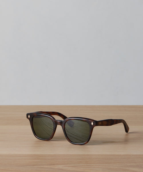 The Broadway Sunglasses by Garrett Leight, featuring dark tortoiseshell frames and green-tinted UV-protective lenses, rest on a light wooden surface with a plain light gray background.